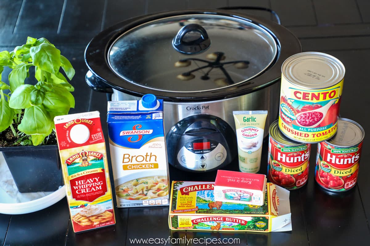 Ingredients for making tomato basil soup in slow cooker including canned tomatoes, chicken broth, cream, basil, butter, garlic