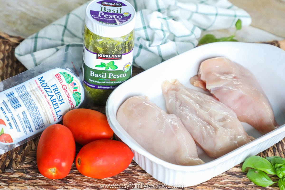 Ingredients for baked caprese chicken including chicken breast, tomatoes, basil pesto, and fresh mozzarella cheese