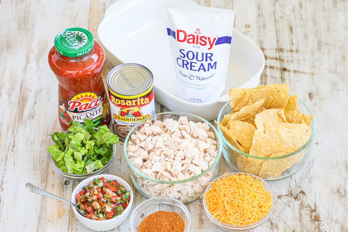 Ingredients for making Chicken Taco Casserole including shredded chicken, refried beans, cheese, salsa, taco seasoning, chips and sour cream