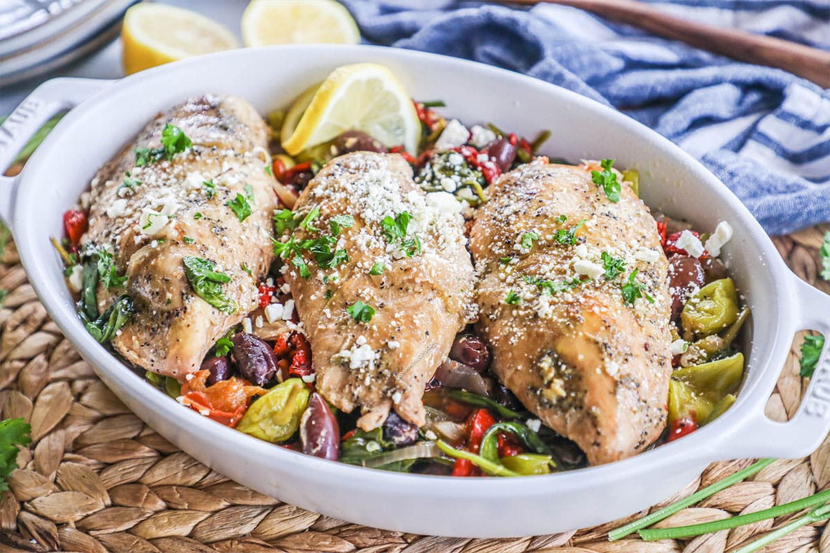 Serving dish with greek chicken with red onion, pepperoncini peppers, roasted red peppers, spinach and olives