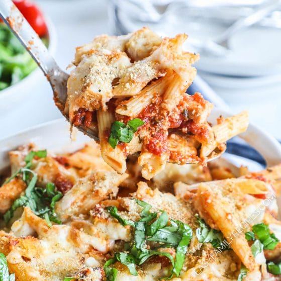 Big spoonful of chicken parmesan casserole with noodles, chicken and pasta sauce covered in cheese and garnished with fresh basil being scooped from casserole dish