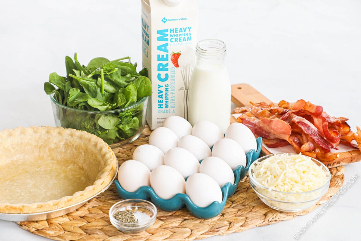 ingredients for spinach bacon quiche that includes a deep dish pie crust, a bowl of fresh spinach, carafe of milk and carton of heavy cream, cooked sliced bacon, bowl of parmesan cheese, salt and pepper.