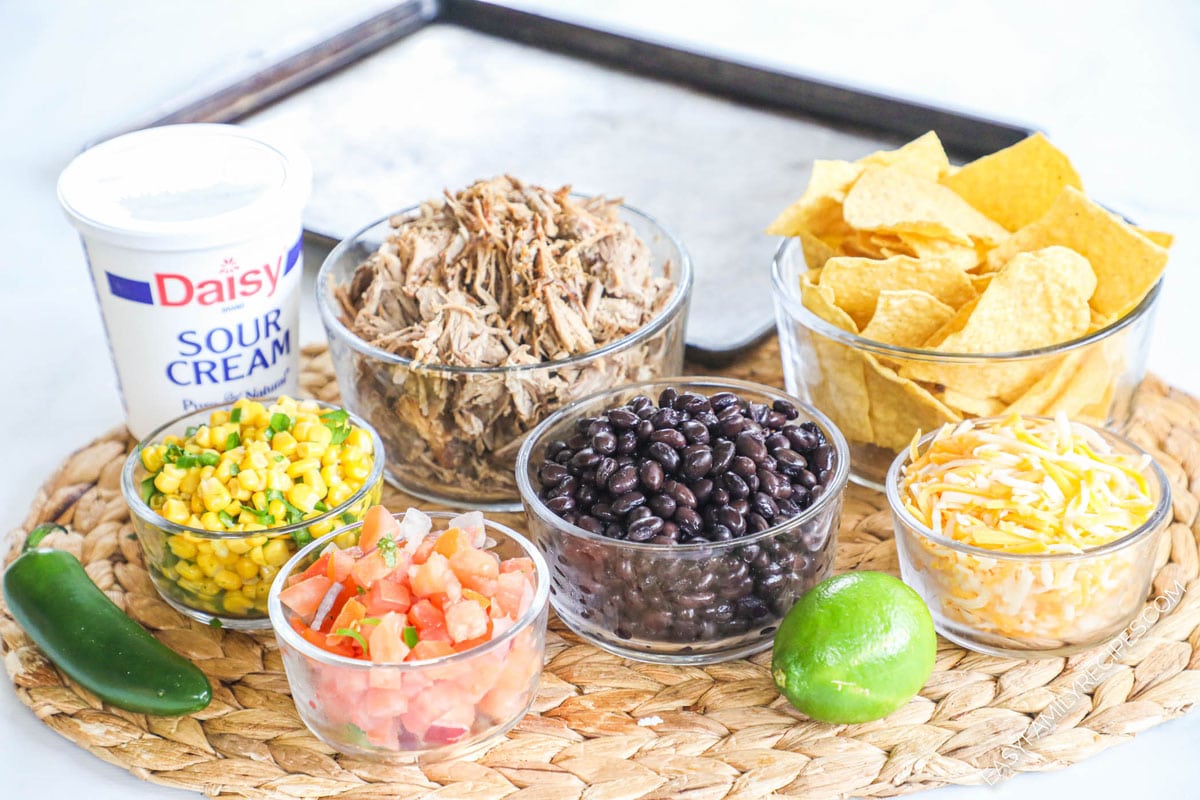 ingredients for recipe: shredded pork carnitas, tortilla chips, shredded cheese, black beans, corn salsa, sour cream, jalepeno, and lime with sheet pan for baking.