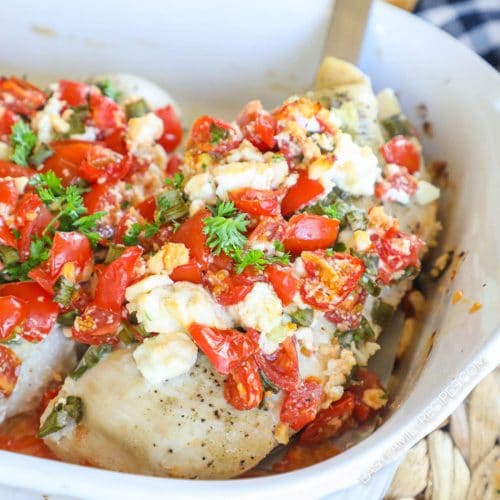 Chicken breast in pan being scooped out. Chicken breast is topped with feta, tomato, and green onions.