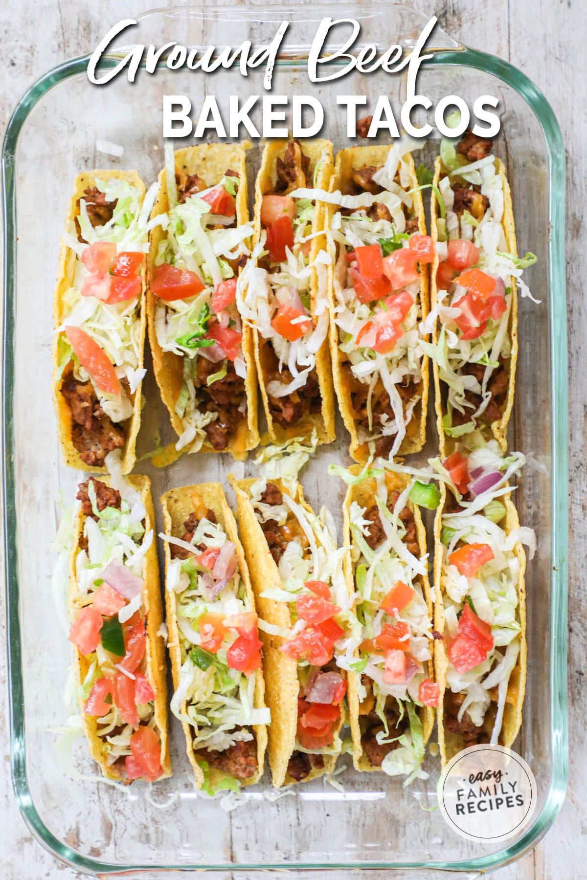 A clear baking dish filled with ten baked crispy ground beef tacos that are all topped with cheese shredded lettuce and diced tomatoes.