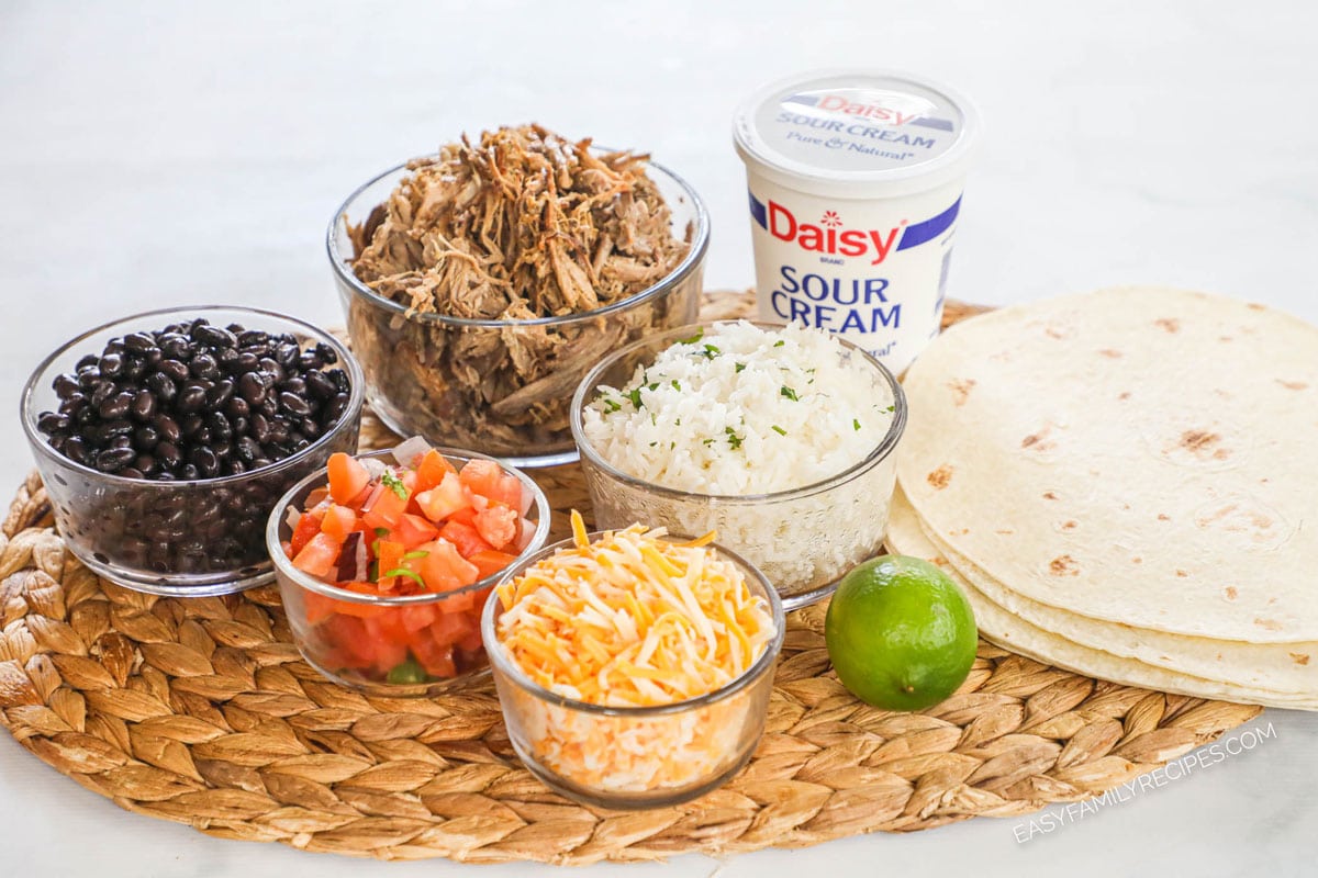 Ingredients for burrito recipe: black beans, tomatoes, shredded cheese, carnitas, rice, sour cream, lime, and large flour tortillas.