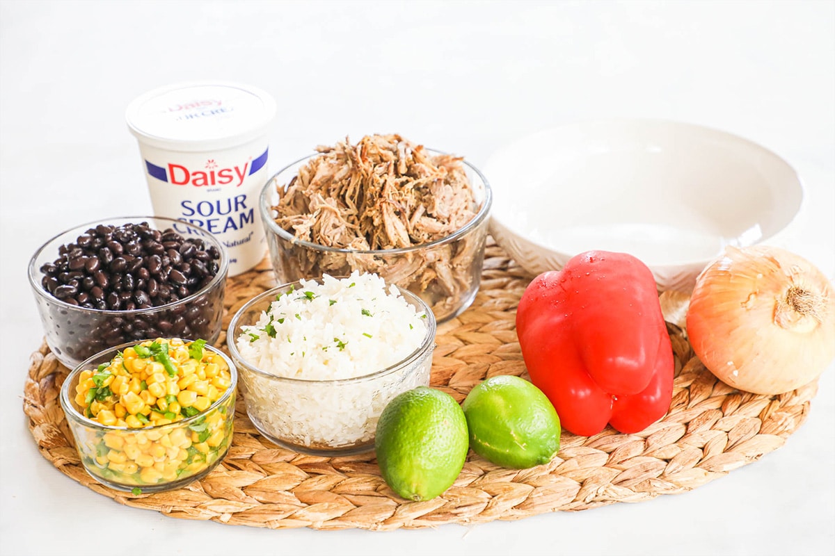 Ingredients for burrito bowl recipe: black beans, red bell pepper, onion, shredded cheese, corn, carnitas, rice, sour cream, and lime