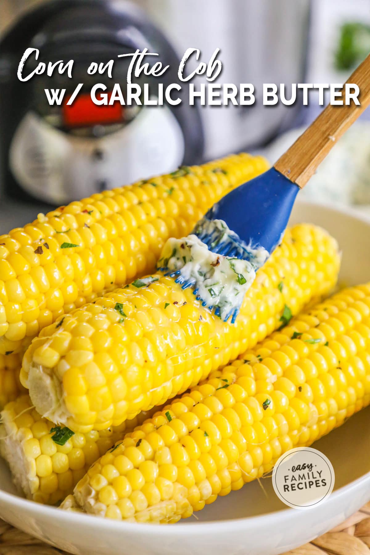 Pile of cooked corn cobs in bowl with garlic herb butter being coated on with a rubber brush.