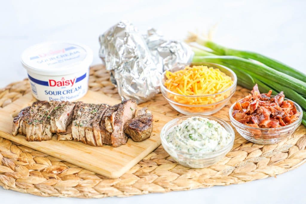 Ingredients displayed to make recipe: sour cream, leftover sliced steak, garlic butter, shredded cheese, bacon pieces, baked potatoes in foil, and green onions.