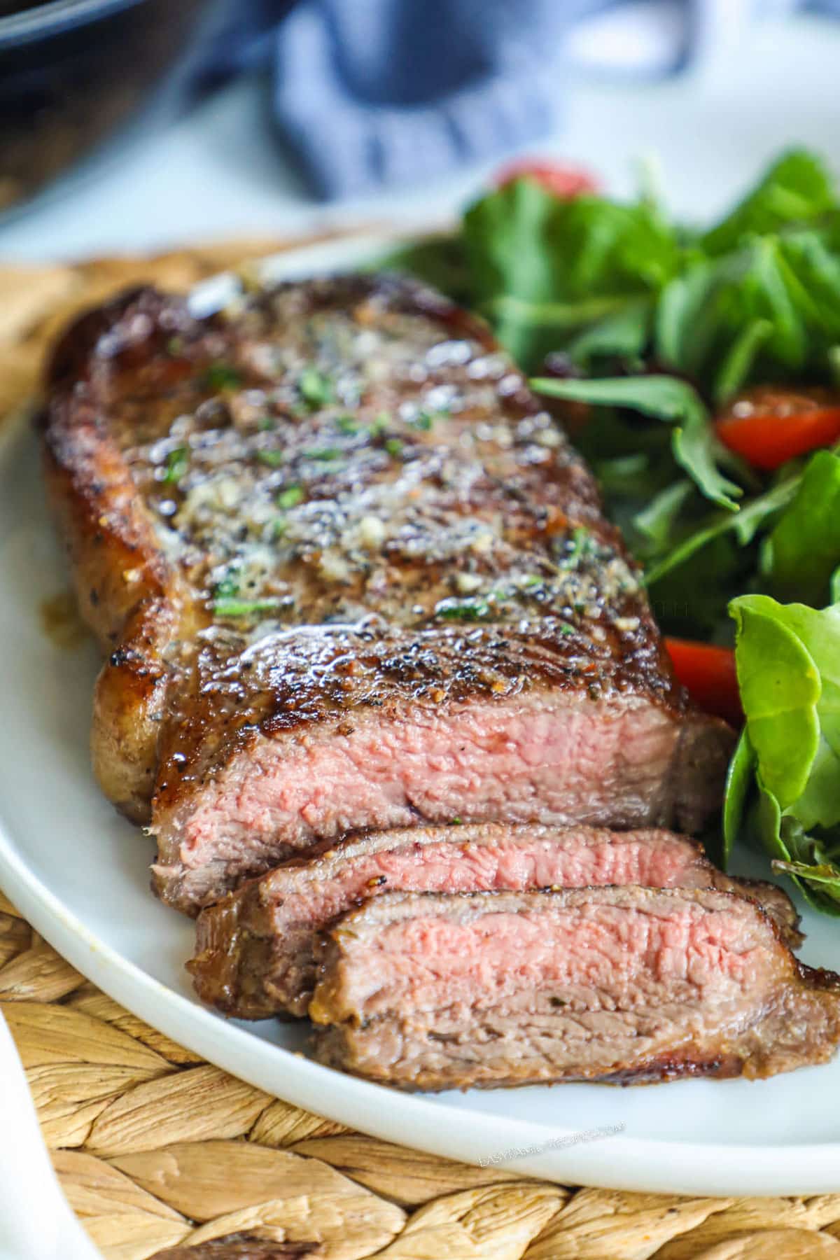 A piece of seared steak sliced on a plate with green herbs on top and served with a side green salad.