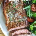 A piece of seared steak sliced on a plate with green herbs on top and served with a side green salad.