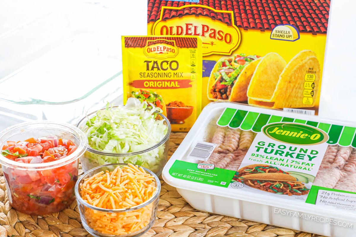 Ingredients to make baked ground turkey tacos including ground turkey, crunchy taco shells, taco seasoning, salsa, and toppings of lettuce and tomato.