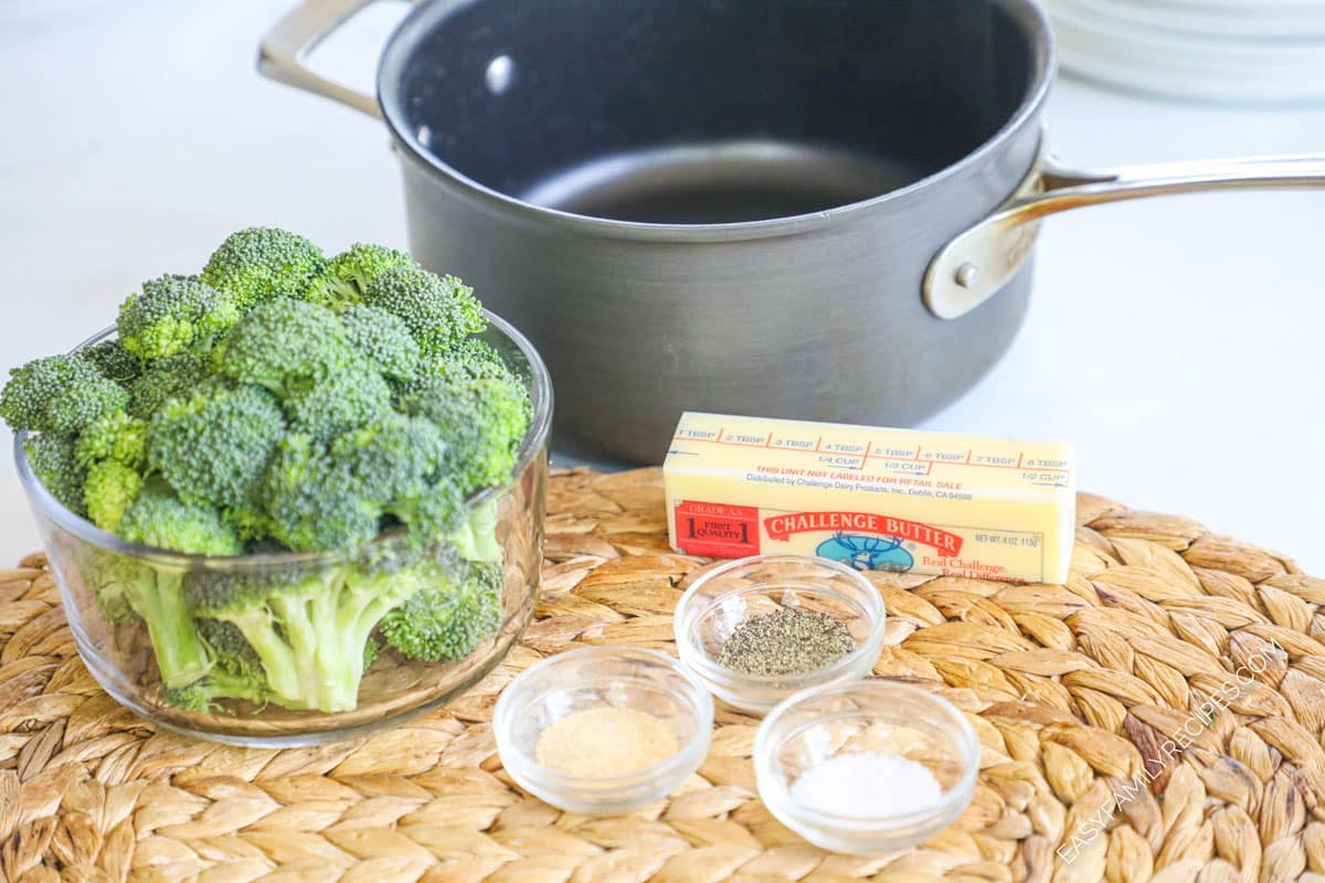 Ingredients for recipe, fresh broccoli florets, garlic powder, pepper, salt, and a stick of butter with a saucepan in the background.