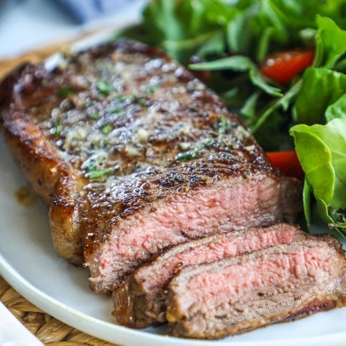 A piece of seared steak sliced on a plate with green herbs on top and served with a side green salad.