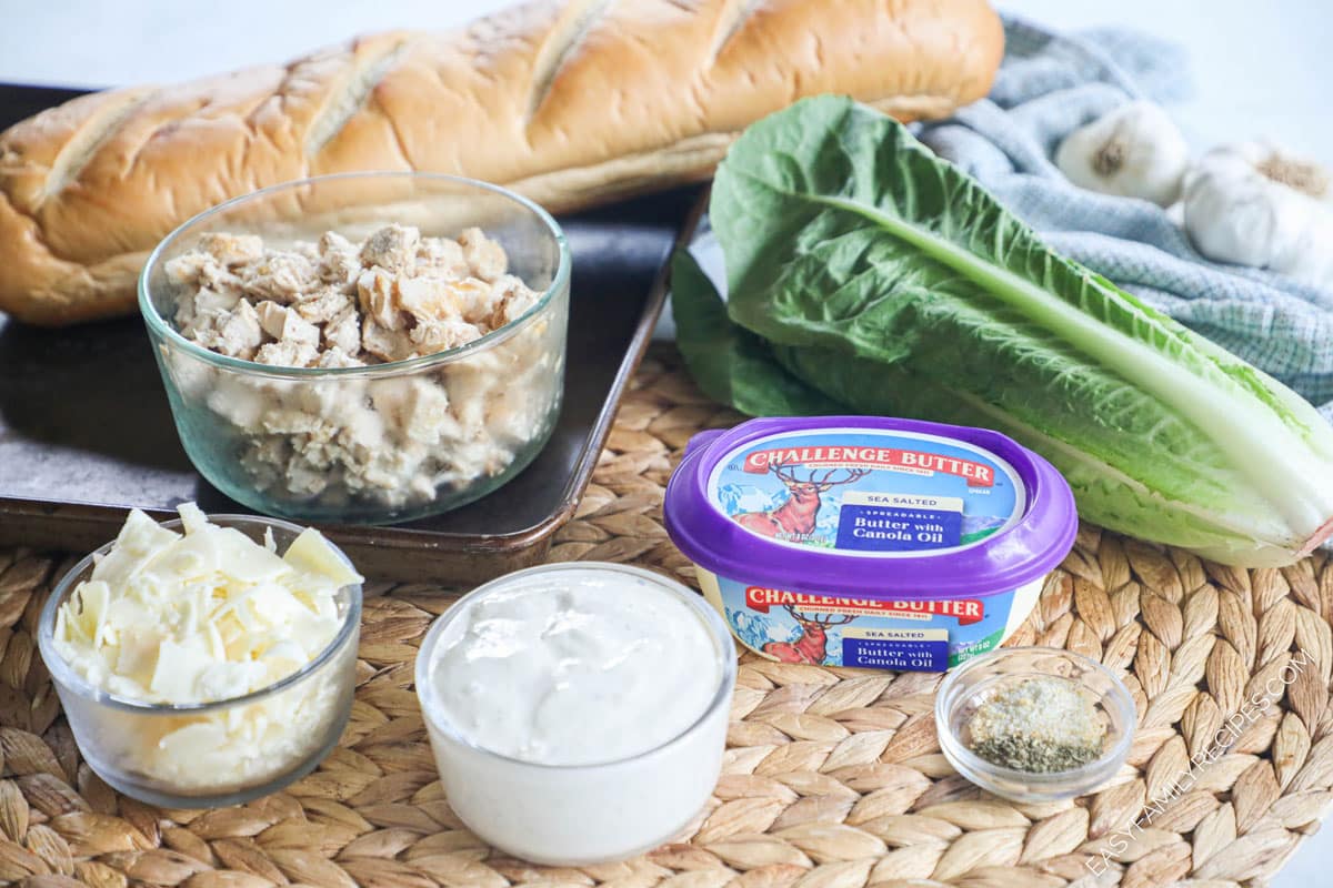 ingredients for making chicken caesar sandwich including french bread, romaine lettuce, parmesan cheese, shredded chicken, creamy caesar dressing.