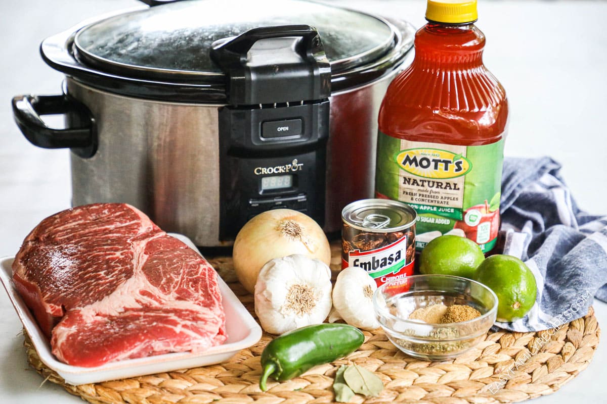 ingredients to make beef barbacoa in a slow cooker including apple juice, chipotle peppers, onion, and lime.