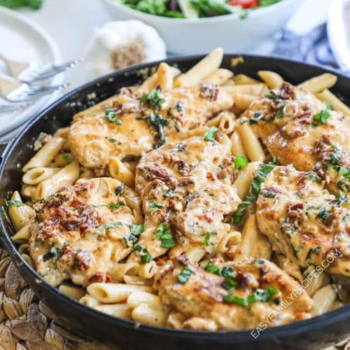 Pasta dish in a skillet with chicken on top and sun dried tomato and basil cream sauce.