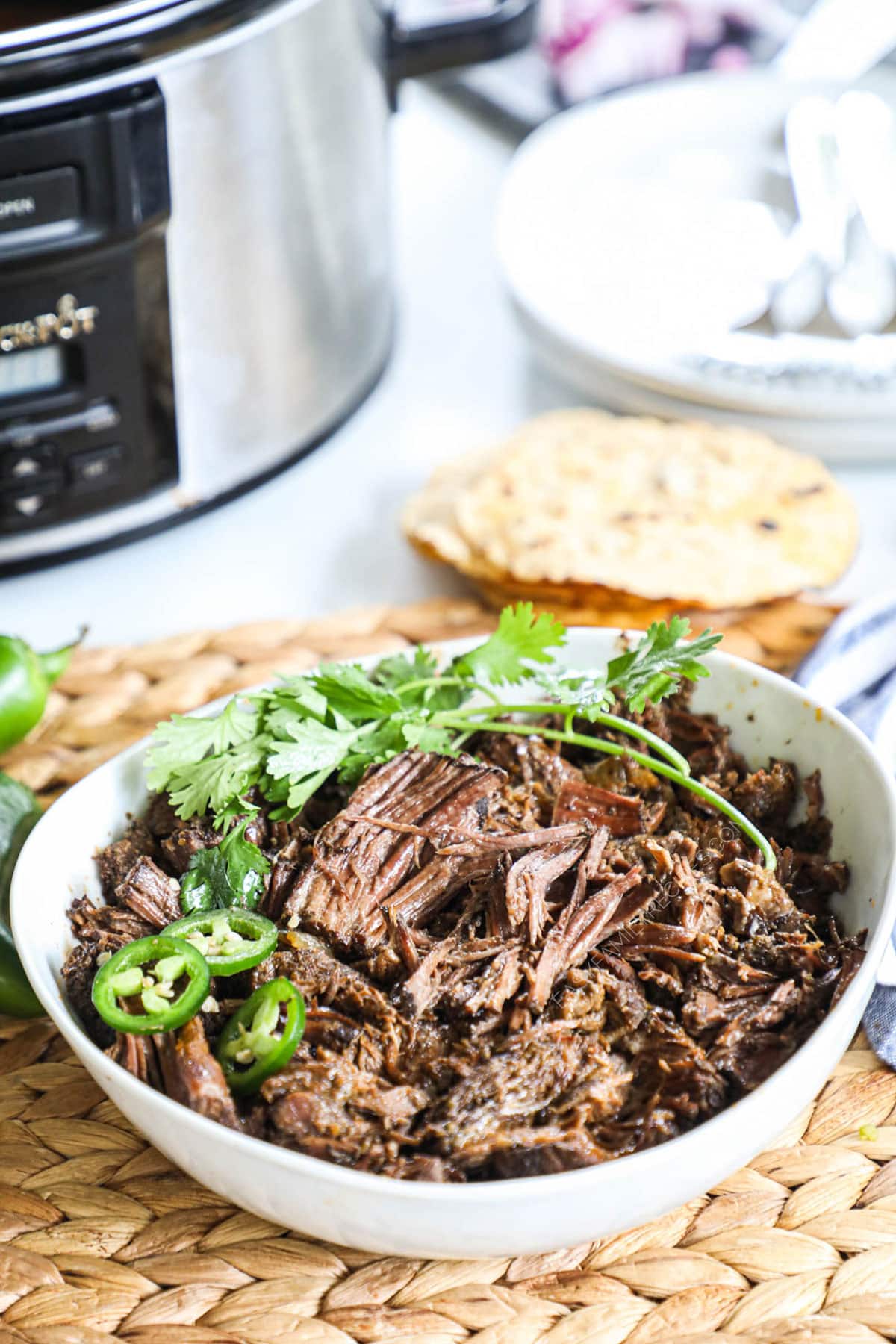 Beef barbacoa in a bowl with cilantro and jalapenos next to tortilla shells and a slow cooker
