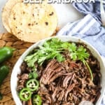 Beef barbacoa in a bowl with cilantro and jalapenos next to tortilla shells