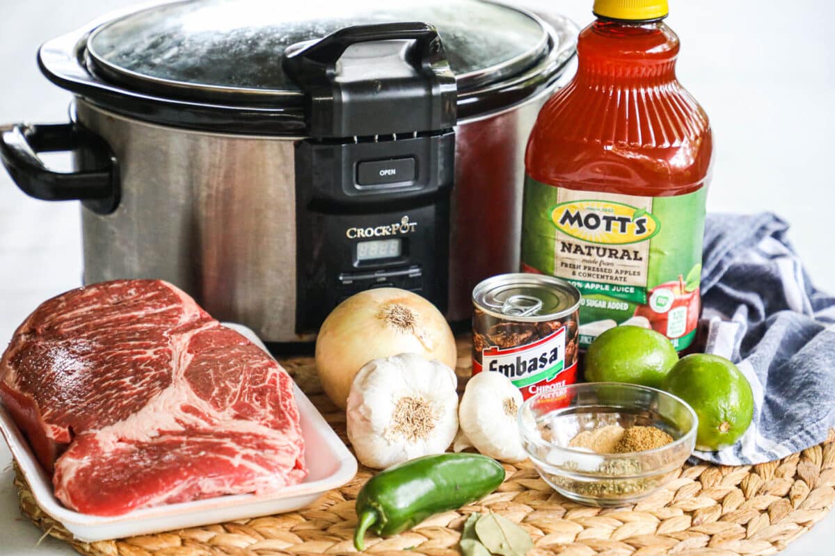 ingredients to make beef barbacoa in a slow cooker including apple juice, chipotle peppers, onion, and lime.