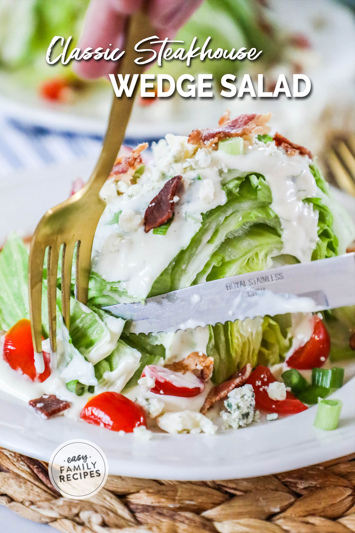 Fork and knife cutting into wedge salad topped with dressing, tomatoes, bacon, and blue cheese on large white plate with another serving behind.
