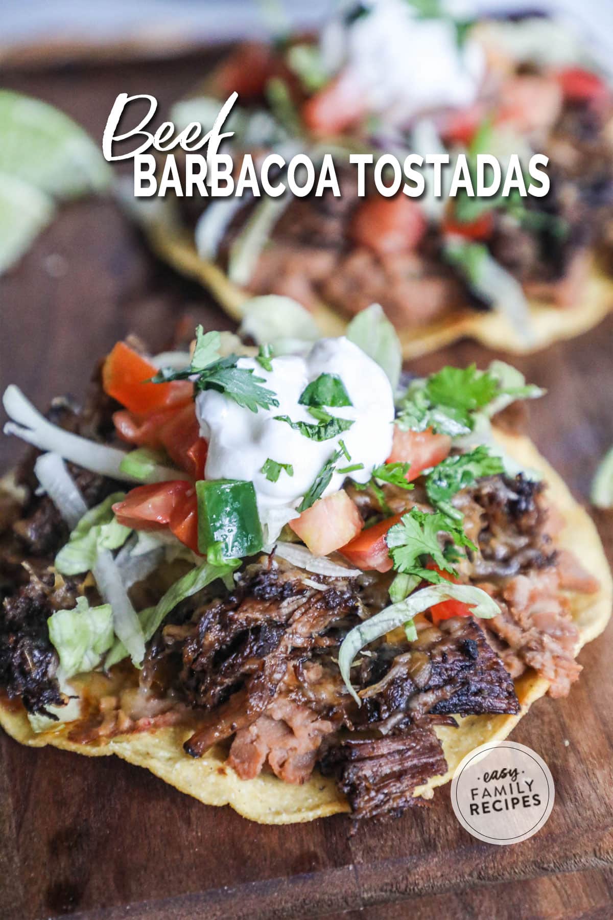 a cutting board topped with a barbacoa tostada garnished with lettuce, tomato, sour cream, and cilantro.