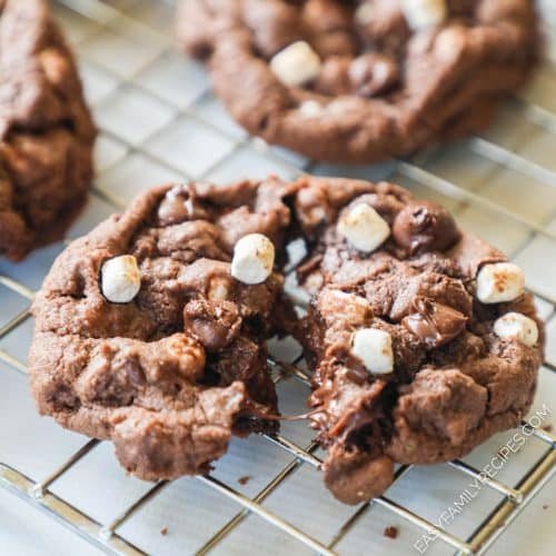 A close up image of a hot chocolate cookie with mini marshmallows on top broken open slightly to show the gooey texture.