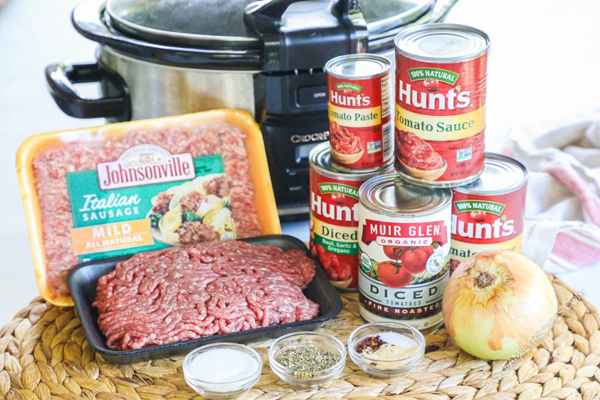 Ingredients for recipe in original packaging: ground Italian mild sausage, ground beef, can tomato paste, 2 cans tomato sauce, can italian diced tomatoes, can fire roasted diced tomatoes, sugar, dried herbs, other spices, and a whole onion sitting infront of Crock pot.