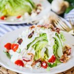 Large wedge of iceberg lettuce topped with creamy dressing, small halved tomatoes, crispy bacon, and blue cheese crumbles.