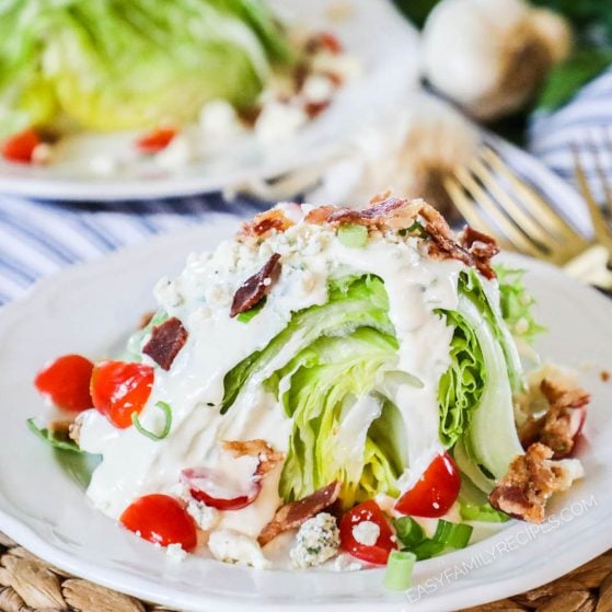 Large wedge of iceberg lettuce topped with creamy dressing, small halved tomatoes, crispy bacon, and blue cheese crumbles.