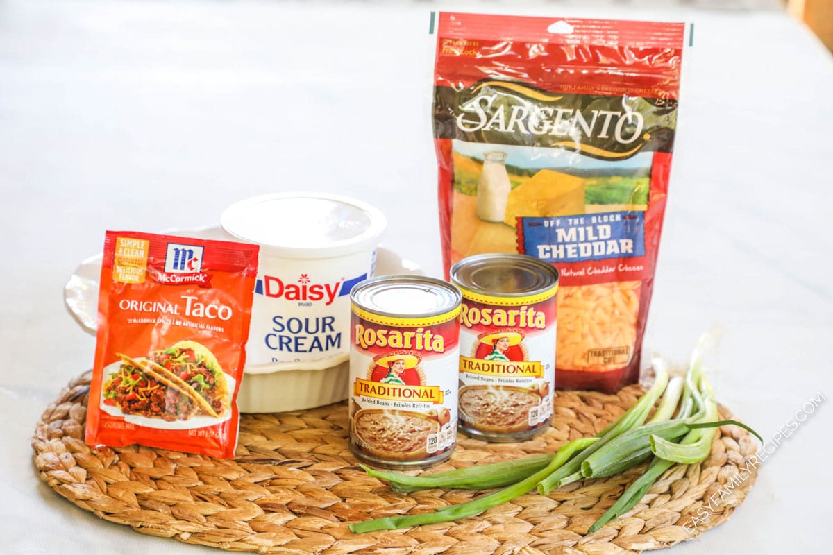 Ingredients for cheesy bean dip including sharp cheddar, sour cream, taco seasoning, refried beans, and green onions.