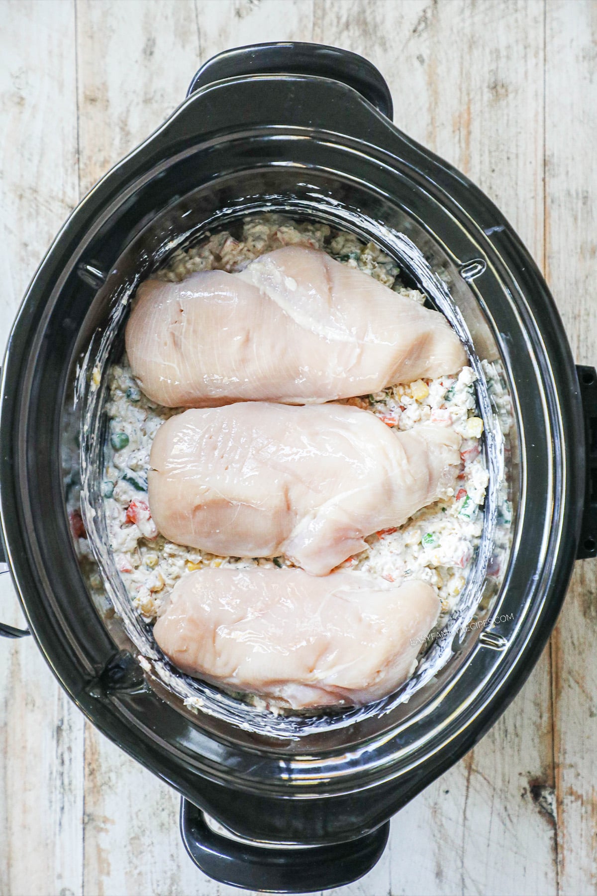 Chicken on top of mixed veggies and other ingredients in a slow cooker before being cooked.