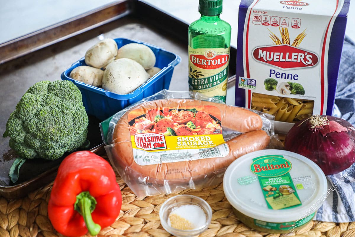 Ingredients for sausage and veggie pesto pasta including smoked sausage, olive oil, mushrooms, bell pepper, broccoli, red onions, pasta, garlic powder, and olive oil.