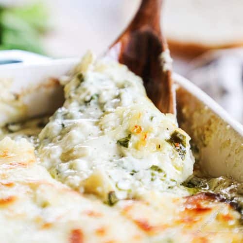 A spoon scooping out gooey spinach artichoke dip from a baking tray.