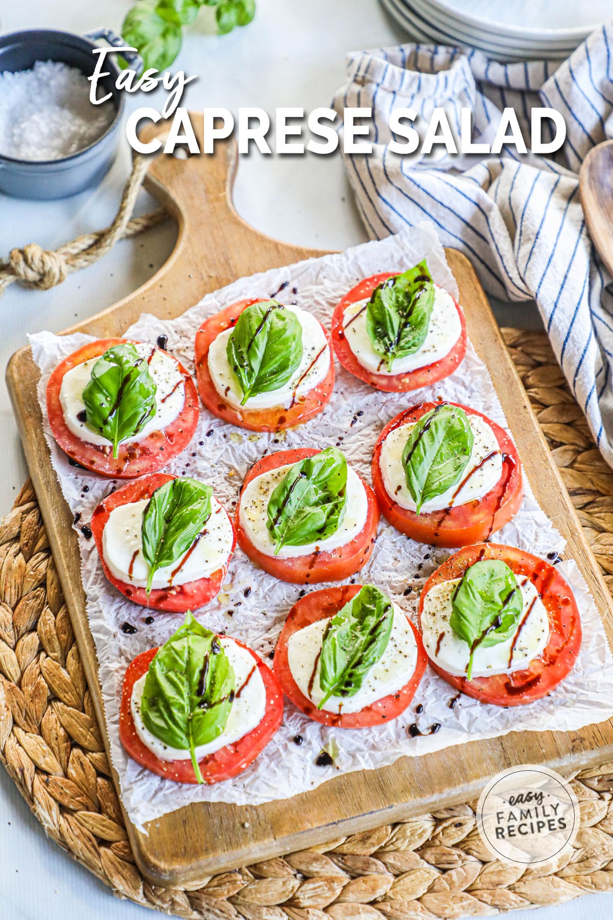 A handled cutting board with a sheet of crinkled parchment paper and caprese salad on top consisting of round slices of tomatoes, sliced cheese, basil, and balsamic glaze.