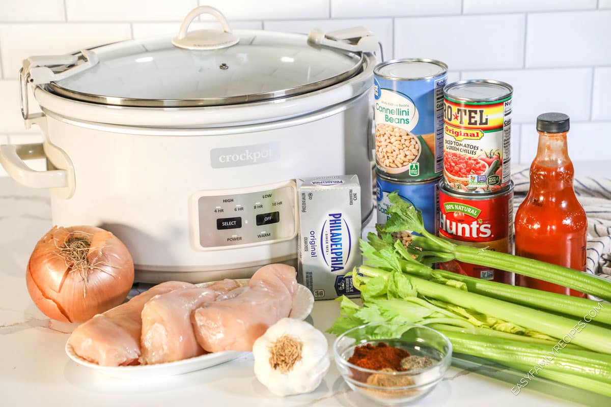 Ingredients for creamy buffalo chicken chili in a crock pot, including chicken, celery, onions, garlic, rotel tomatoes, tomato sauce, and hot sauce