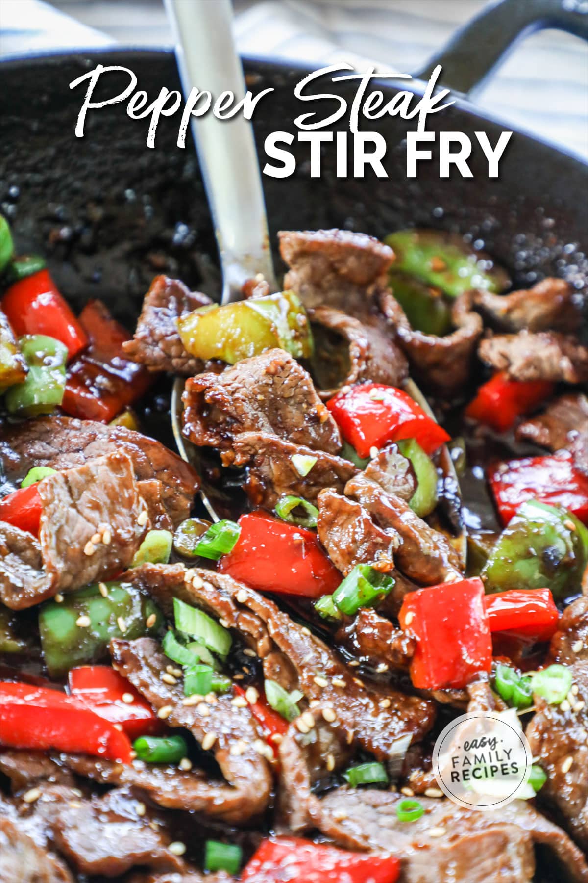 A metal spoon scooping stir fried steak with red and green peppers from a large black pan.
