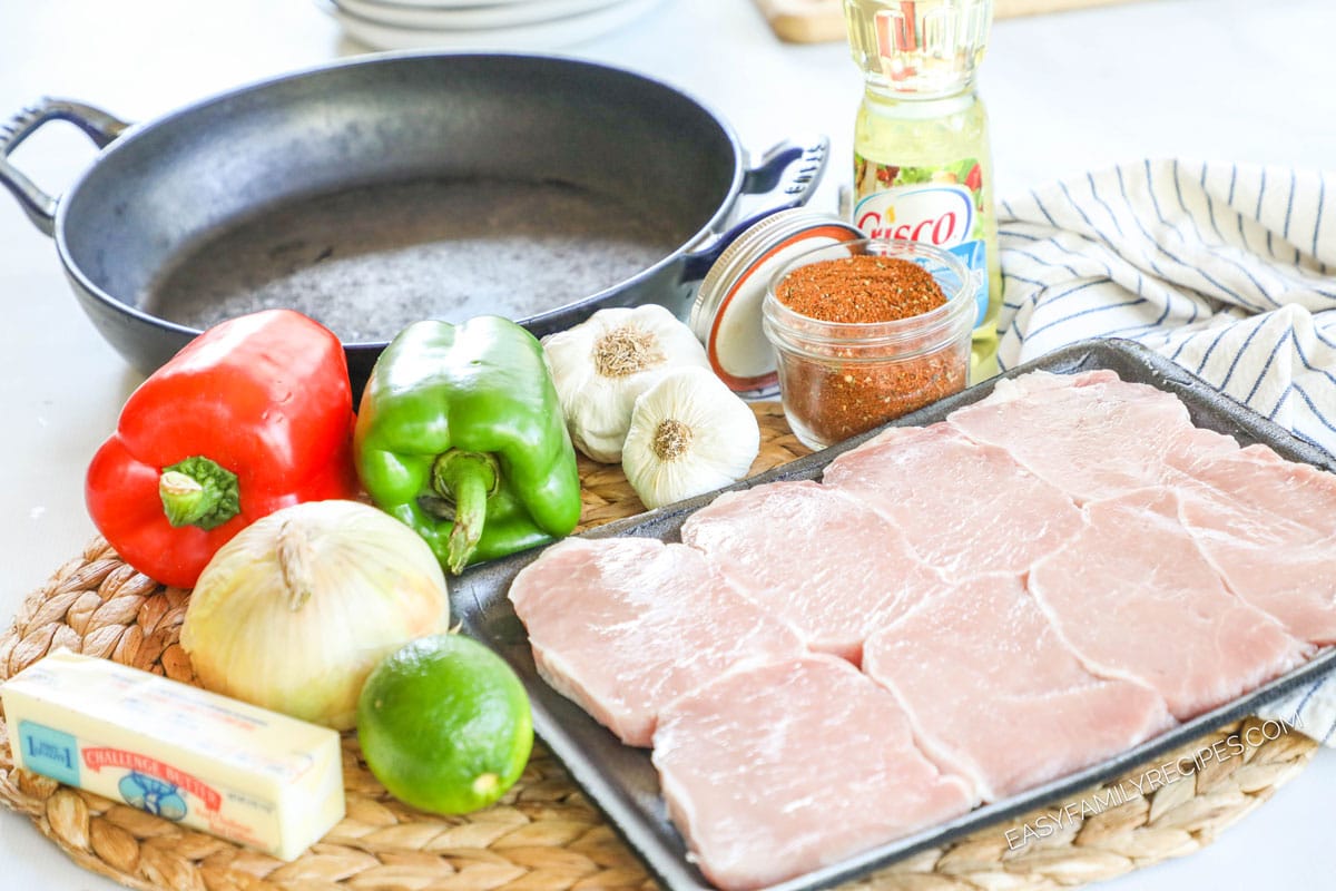 Ingredients for recipe before prepping: red and green bell peppers, onion, butter, lime, garlic heads, fajita seasonings, oil and pork chops with a pan nearby.