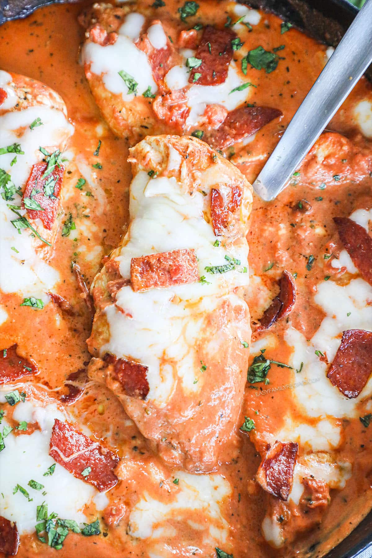 chicken breasts cooked in a skillet with tomato sauce and pepperoni pieces is being scooped out of the dish to serve