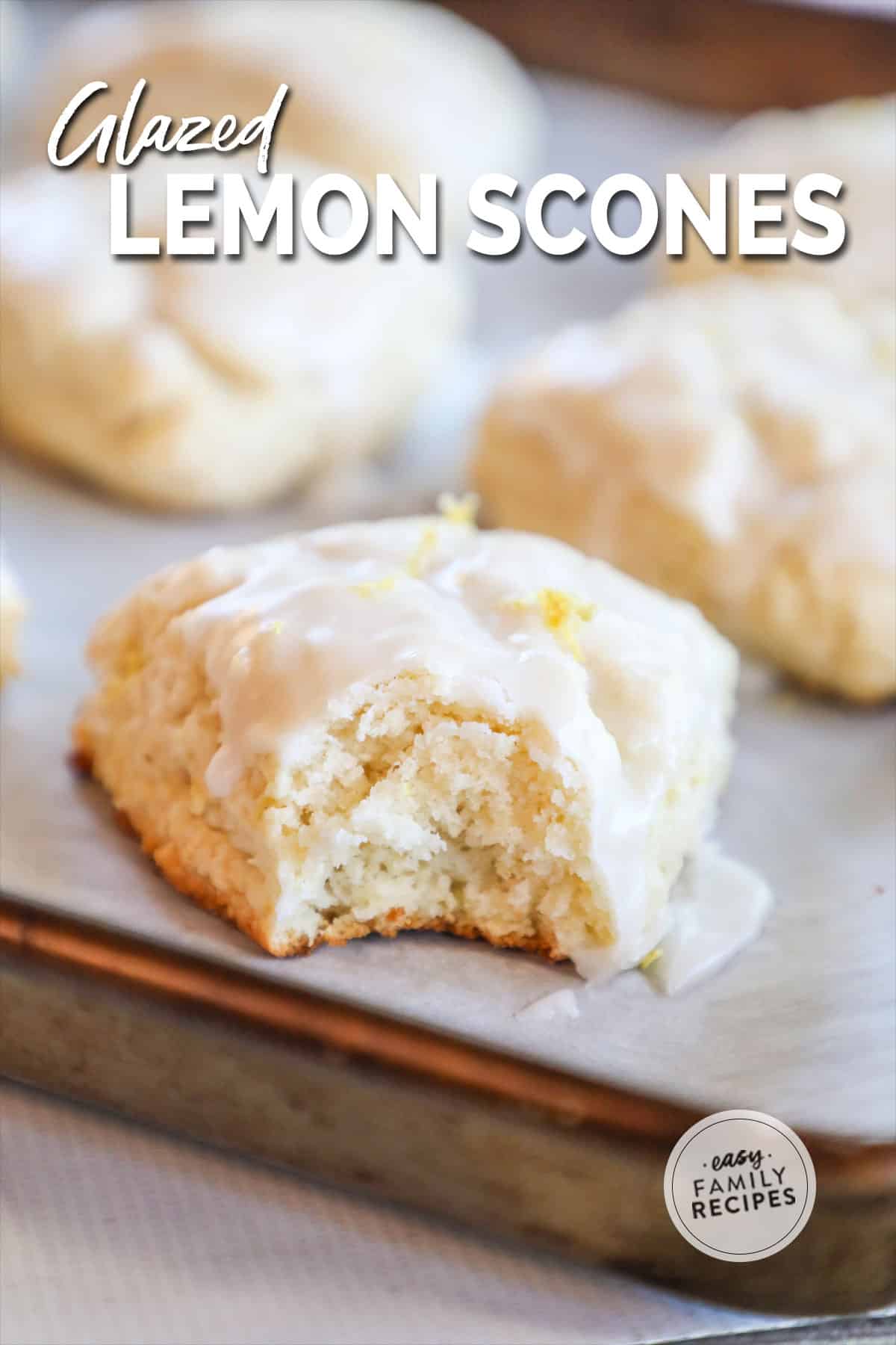 Close up of a glazed lemon scone with a bite taken out on a parchment-lined baking sheet with more scones nearby.