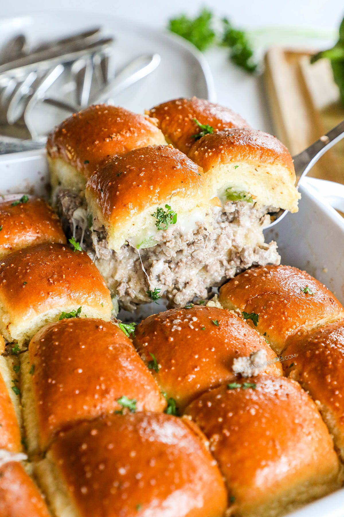 A baking dish full of Philly Cheesesteak Sliders being scooped out with a spoon. The sliders have ground beef, onions and bell peppers