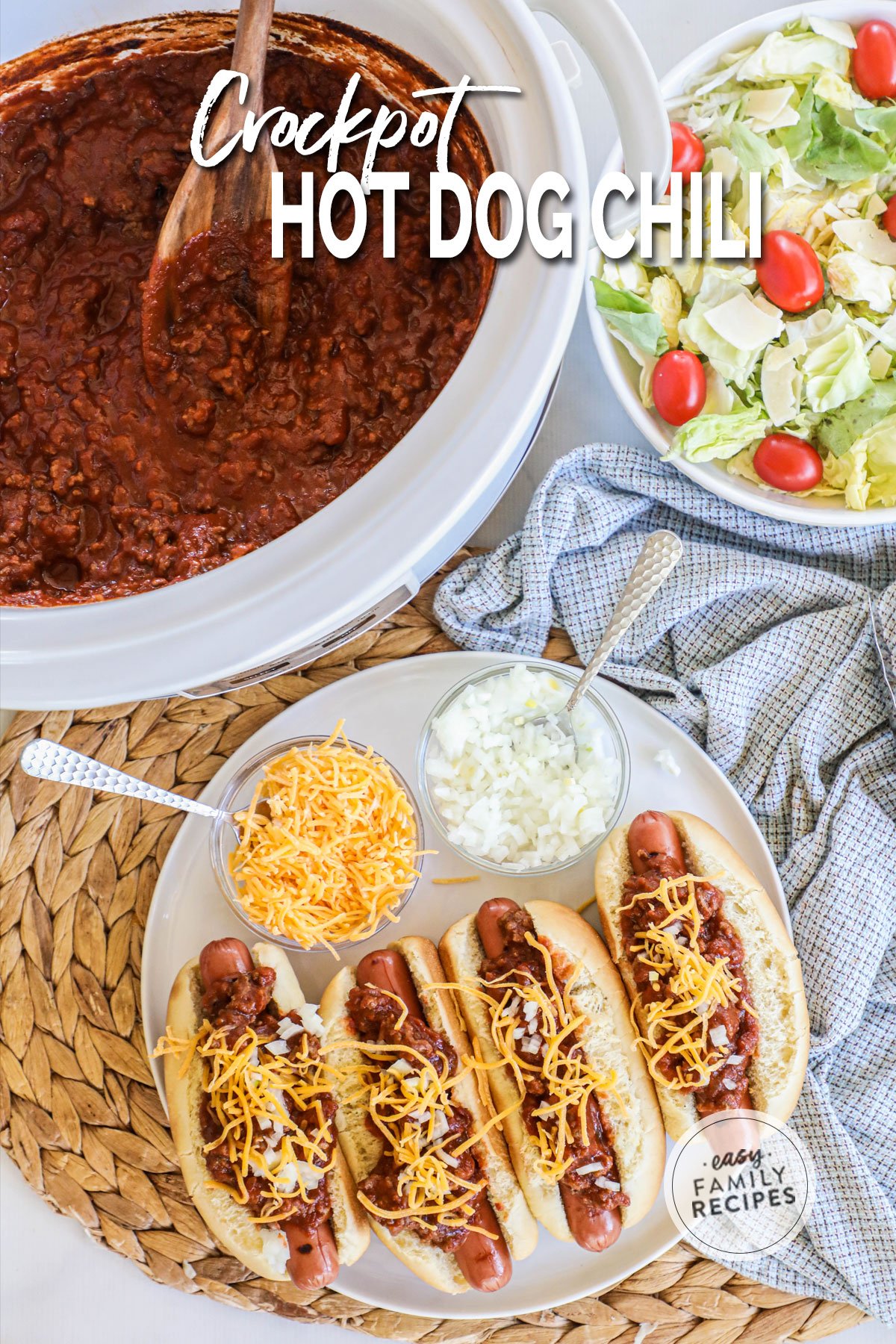 a Crock Pot full of chili next to a salad and a platter of chili dogs with cheese.
