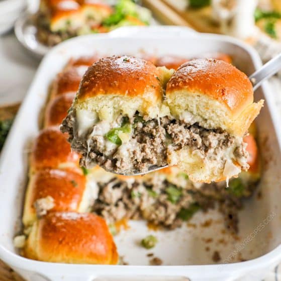 A baking dish full of Philly Cheesesteak Sliders being scooped out with a spoon. The sliders have ground beef, onions and bell peppers