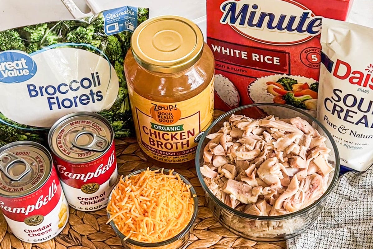 Ingredients for recipe in original packaging and bowls: bag of frozen broccoli florets, can of each cheddar cheese and cream of chicken condensed soups, jar of chicken broth, shredded cheddar cheese, chopped cooked chicken, box of white rice, and sour cream