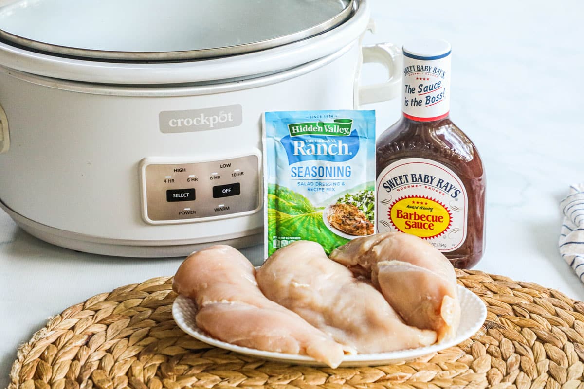 Ingredients for recipe infront of a crock pot: raw chicken breast, ranch seasoning packet, and Sweet Baby Ray's barbeque sauce.