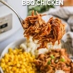 Fork lifting up a bite of Crock Pot BBQ Chicken above a plate with shredded chicken, corn, and mashed potatoes and crockpot nearby.
