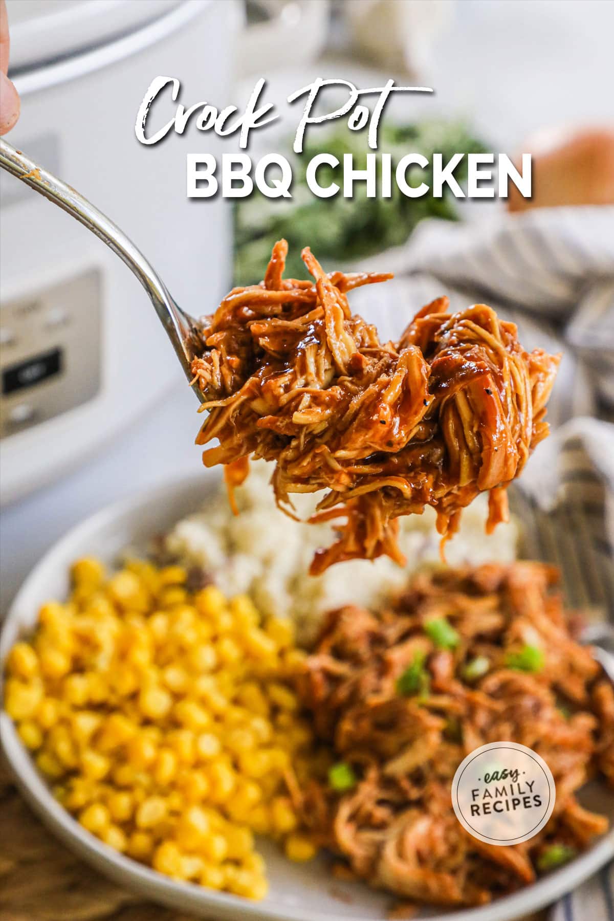 Fork lifting up a bite of Crock Pot BBQ Chicken above a plate with shredded chicken, corn, and mashed potatoes and crockpot nearby.