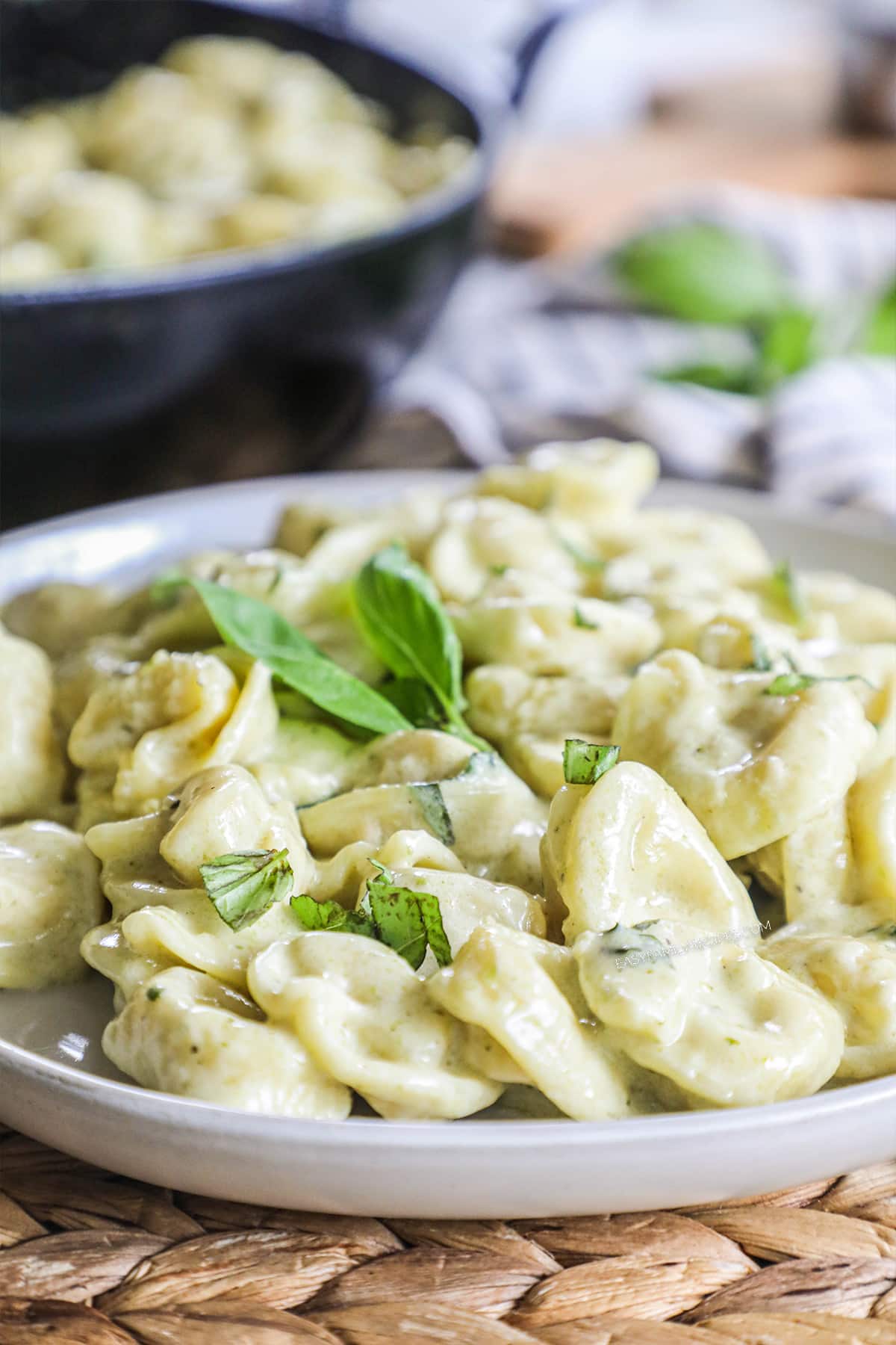 A white plate of tortellini tossed with pesto cream sauce and garnished with fresh basil.