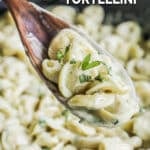a wooden spoon scooping tortellini with pesto cream sauce and fresh basil out of a large, black skillet.