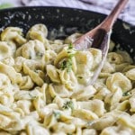 a wooden spoon scooping tortellini with pesto cream sauce and fresh basil out of a large, black skillet.
